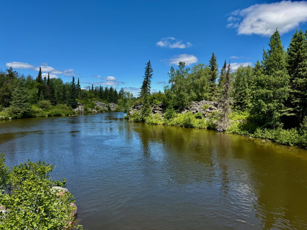 River flowing through a boreal forest landscape with coniferous and deciduous trees, rocky outcrops, and clear blue sky with white clouds