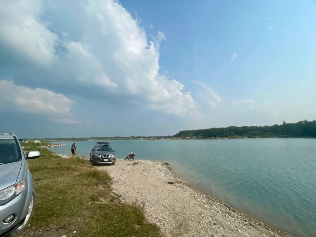 Vehicles parked on a gravel beach beside turquoise water, with people enjoying the shoreline on a sunny day under partly cloudy skies