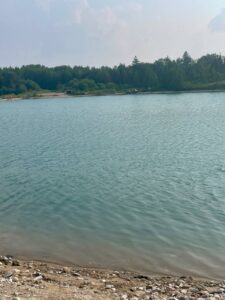 Calm lake with rocky shoreline in foreground, dense coniferous forest along far shore, and small buildings visible near the water's edge