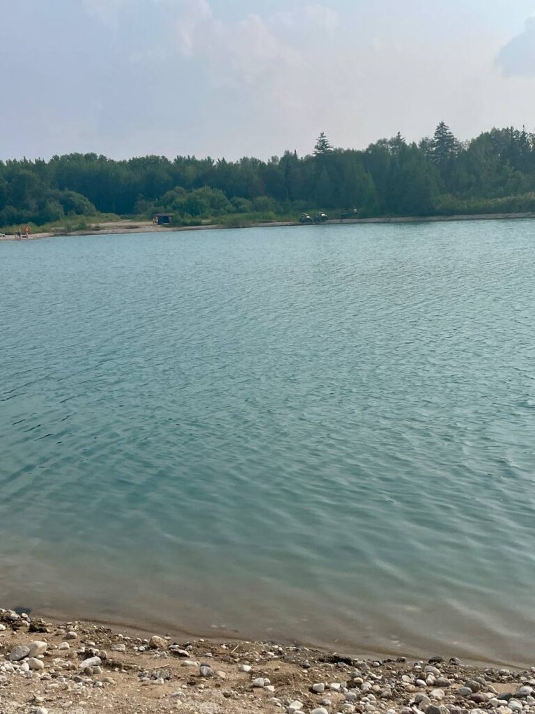 Calm lake with rocky shoreline in foreground, dense coniferous forest along far shore, and small buildings visible near the water's edge