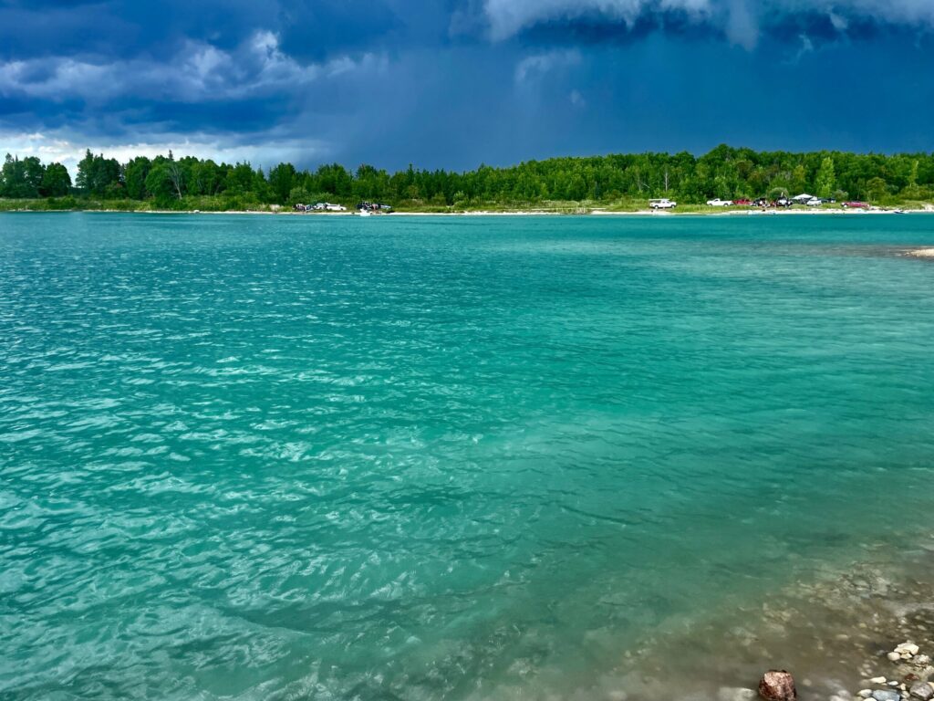 Turquoise water along a sandy beach with forested shoreline, boats, and vehicles visible across the water under a partly cloudy sky