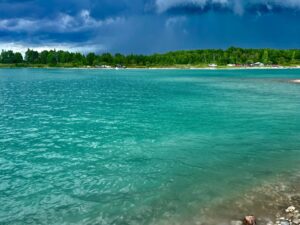 Turquoise water along a sandy beach with forested shoreline, boats, and vehicles visible across the water under a partly cloudy sky