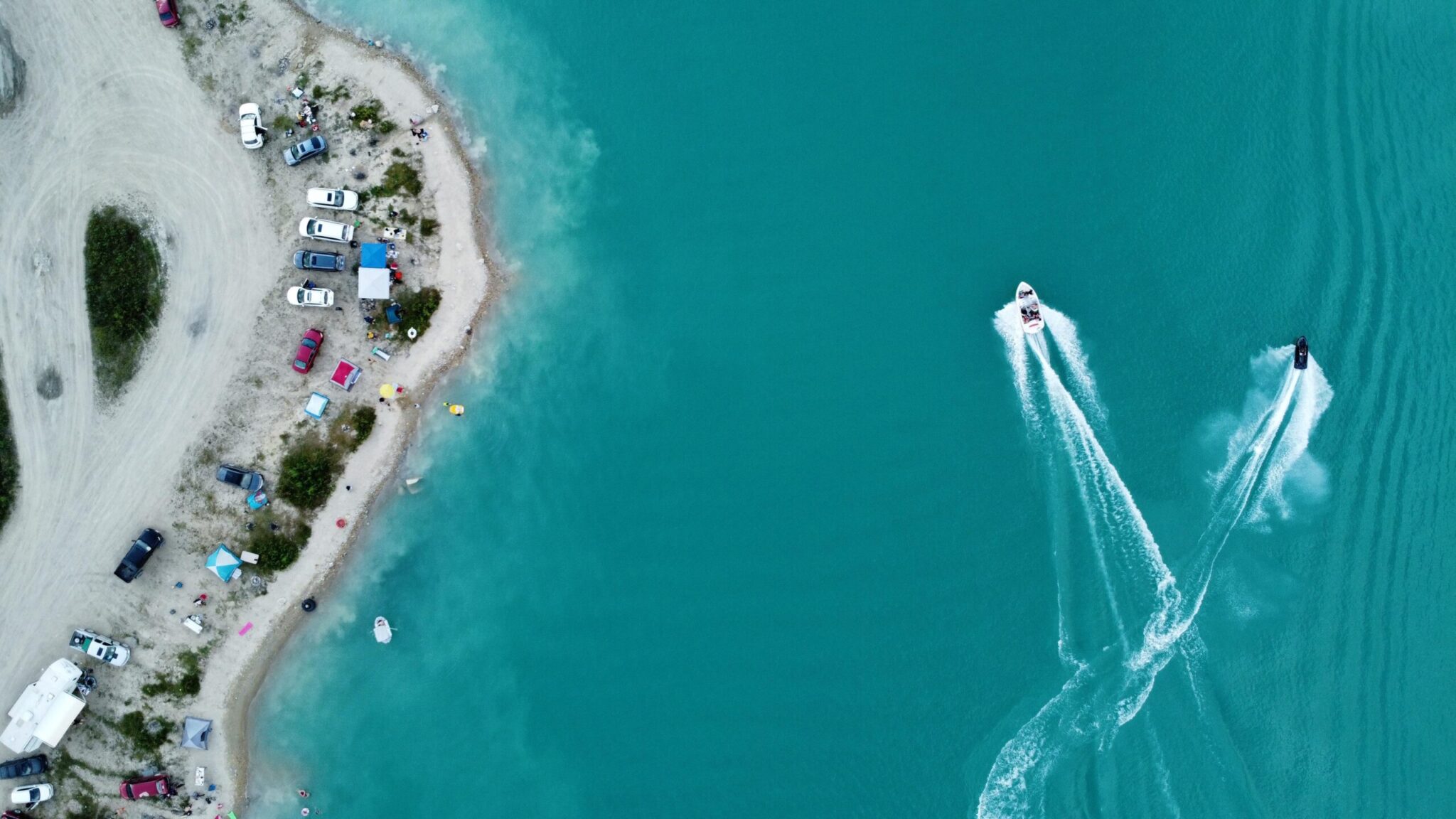 Aerial view of a sandy beach with parked vehicles and beachgoers along the shoreline, and two speedboats creating wake trails in turquoise water offshore