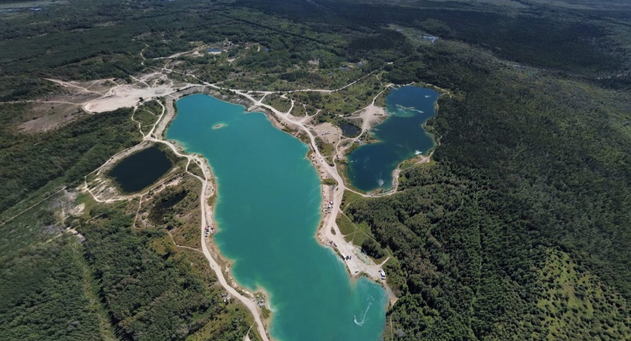 Aerial view of turquoise glacial lakes surrounded by dense coniferous forest, with winding roads and cleared areas connecting multiple lakes in a mountainous region