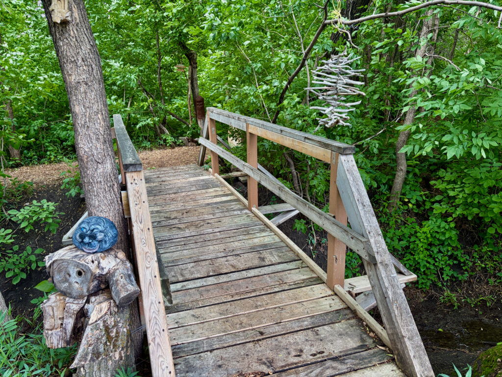 Wooden boardwalk with railings extending through dense green forest, with a carved wooden figure wearing a blue mask at the entrance