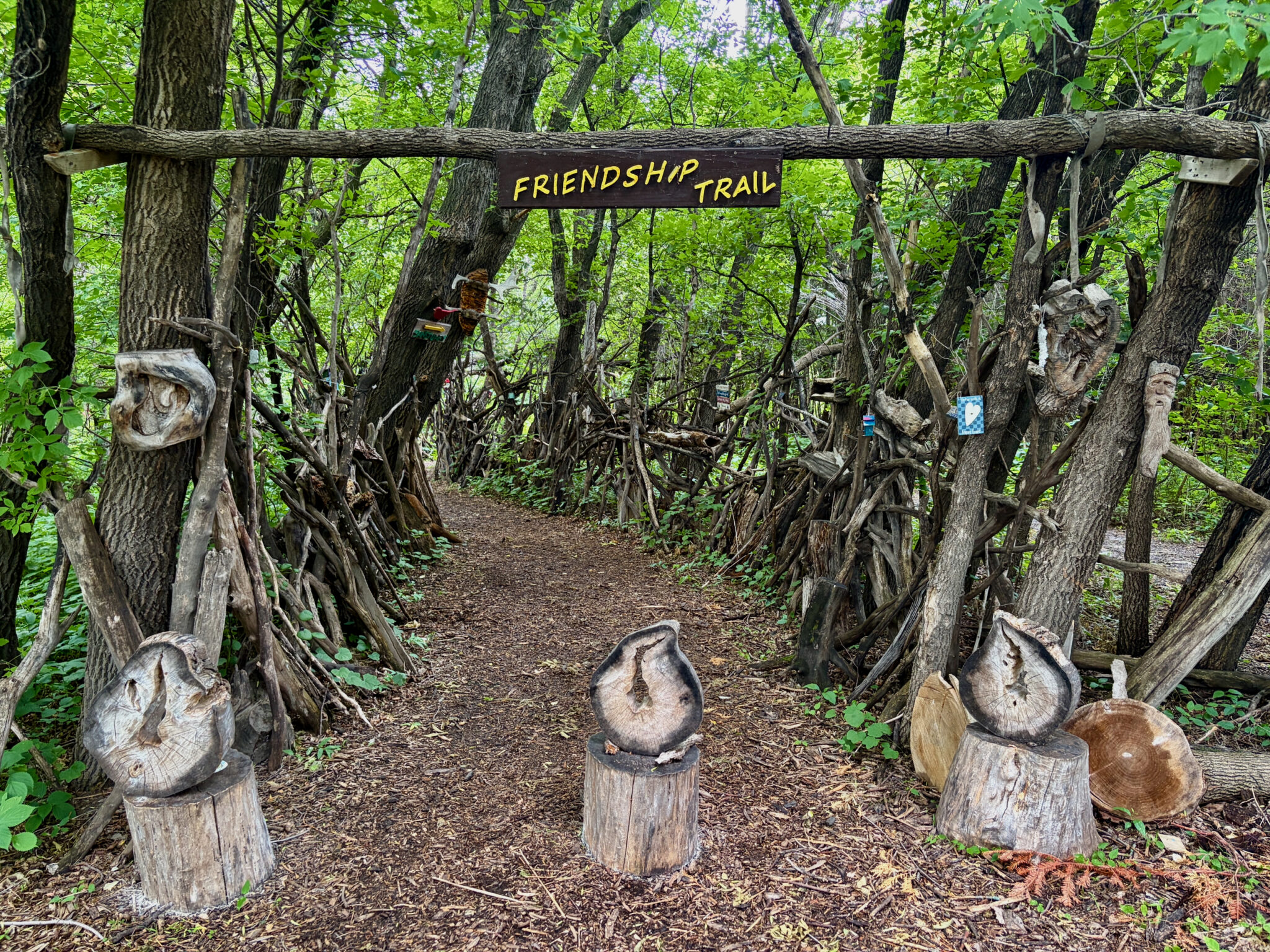 Entrance to Friendship Trail, a forest path lined with wooden carved sculptures and a yellow sign on a fallen log overhead, surrounded by dense green trees