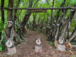 Entrance to Friendship Trail, a forest path lined with wooden carved sculptures and a yellow sign on a fallen log overhead, surrounded by dense green trees