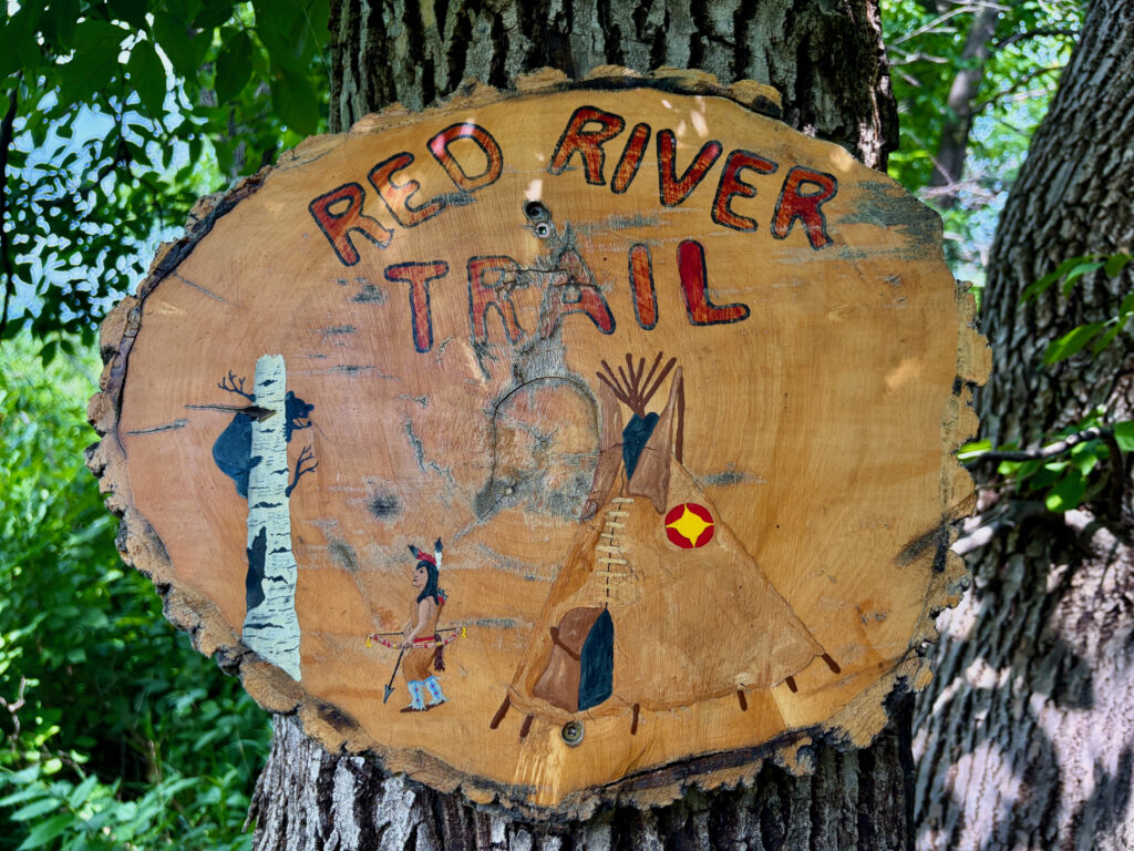 Red River Trail sign painted on tree cross-section showing stylized figures of a birch tree, canoeist, and woodpecker among forest imagery