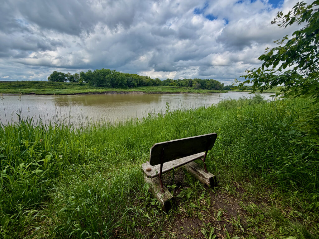 Wooden bench overlooking a river with green marshland, trees lining the far bank, and cloudy sky above