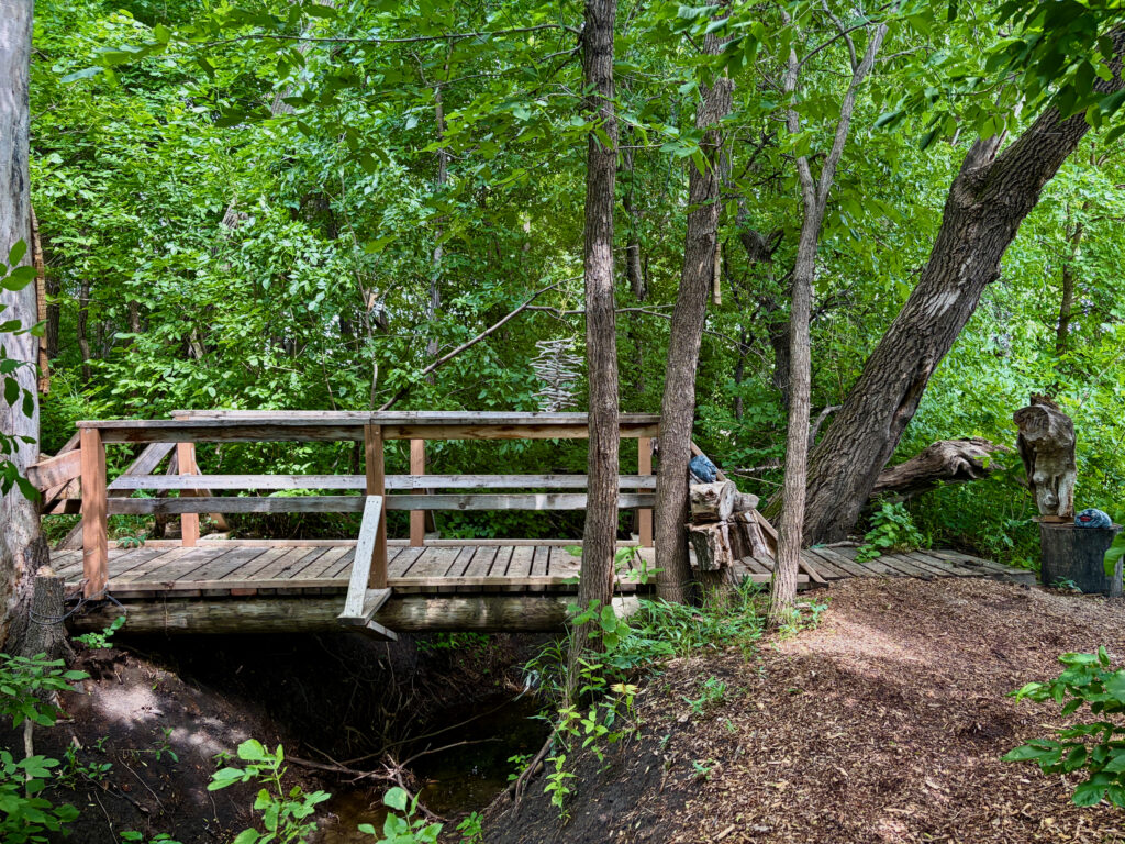 Wooden boardwalk with railings extending through dense green forest, with a carved wooden figure wearing a blue mask at the entrance