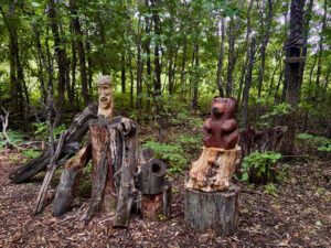 Wooden carved sculptures of a human head and a bear displayed on tree stumps in a dense forest