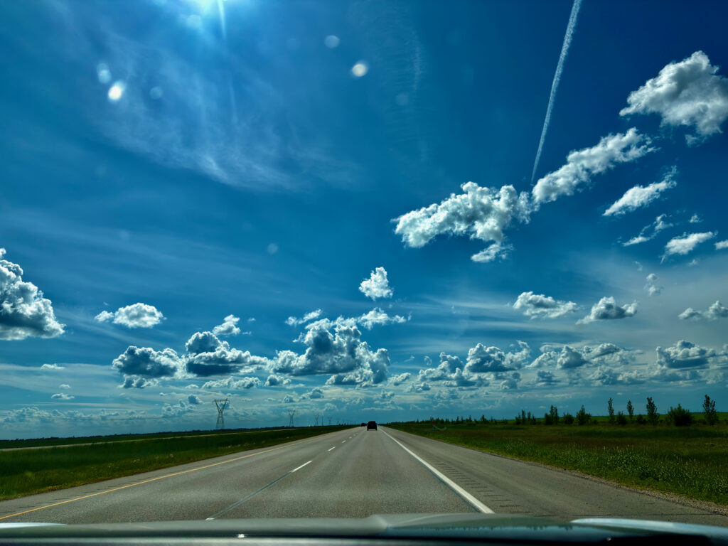 Straight highway stretching across flat prairie landscape with green fields on both sides, power lines along the horizon, and a bright blue sky with white cumulus clouds and a contrail