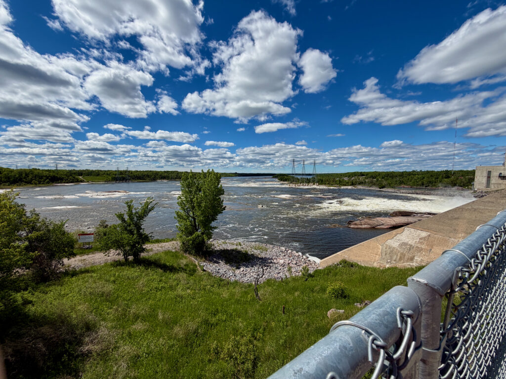 View of a wide river with turbulent waters flowing past forested banks, transmission towers visible in the distance, concrete dam structure and metal railing in foreground under a blue sky with white clouds