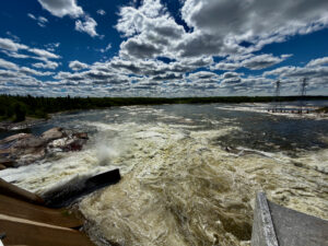 Turbulent river with whitecaps and logs in foreground, forested shoreline, power transmission towers, and blue sky with white clouds