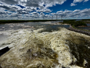 Turbulent river with high water levels and whitecaps flowing past three electrical transmission towers on the far bank, surrounded by forested shoreline under a partly cloudy sky