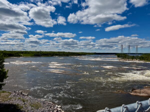 Wide river with turbulent waters flowing past forested shorelines under a blue sky with white clouds, electrical transmission towers visible on the far bank
