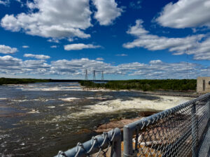 Rushing river with transmission towers and forested shoreline under blue sky with white clouds, viewed from behind a metal fence