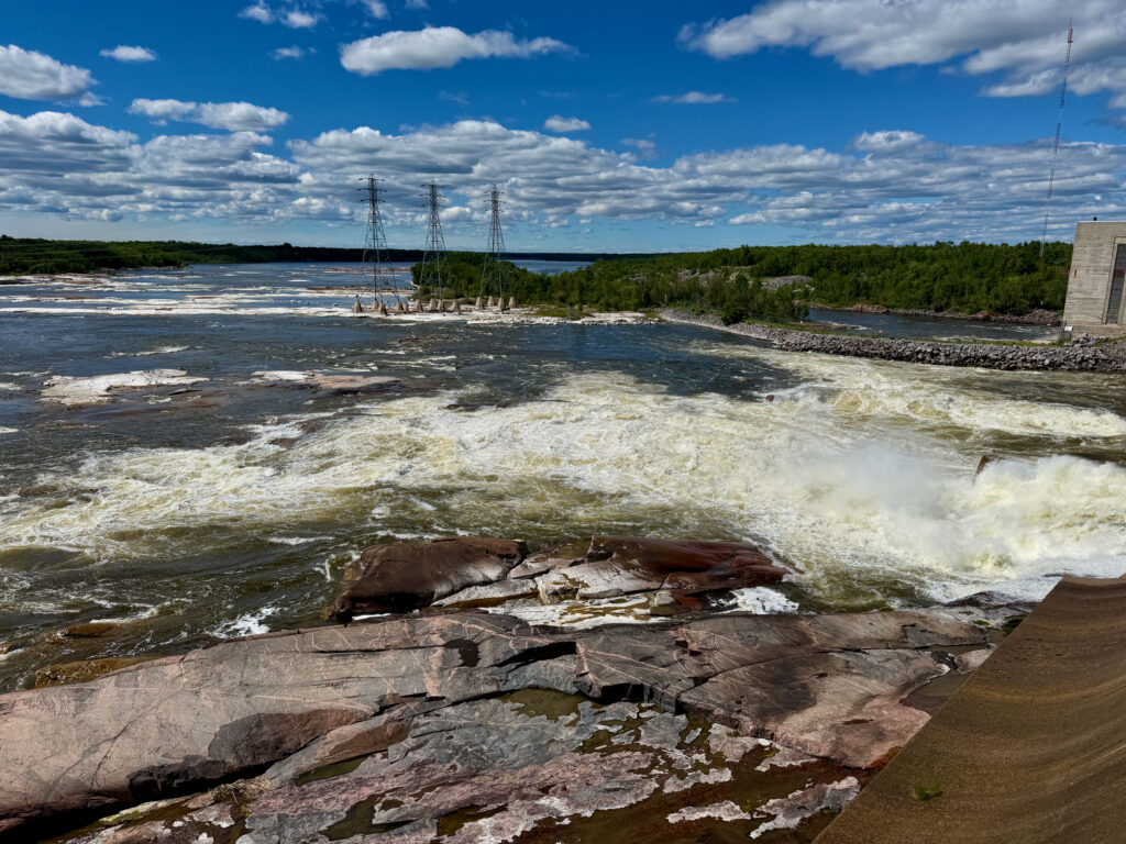 Turbulent river with rocky shoreline, driftwood, electrical transmission towers, forested banks, and a concrete structure on the right under a partly cloudy sky
