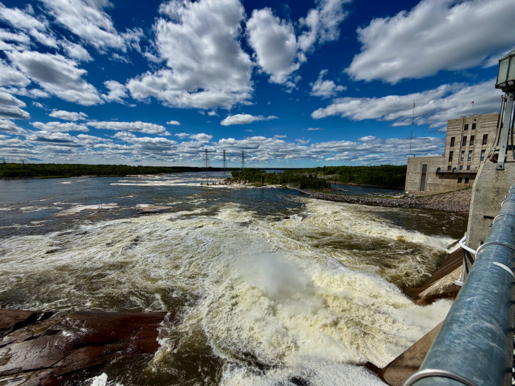 Turbulent water cascades from a dam spillway into a river, with forested shoreline and transmission towers visible under a blue sky with white clouds