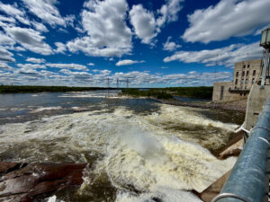 Turbulent water cascades from a dam spillway into a river, with forested shoreline and transmission towers visible under a blue sky with white clouds