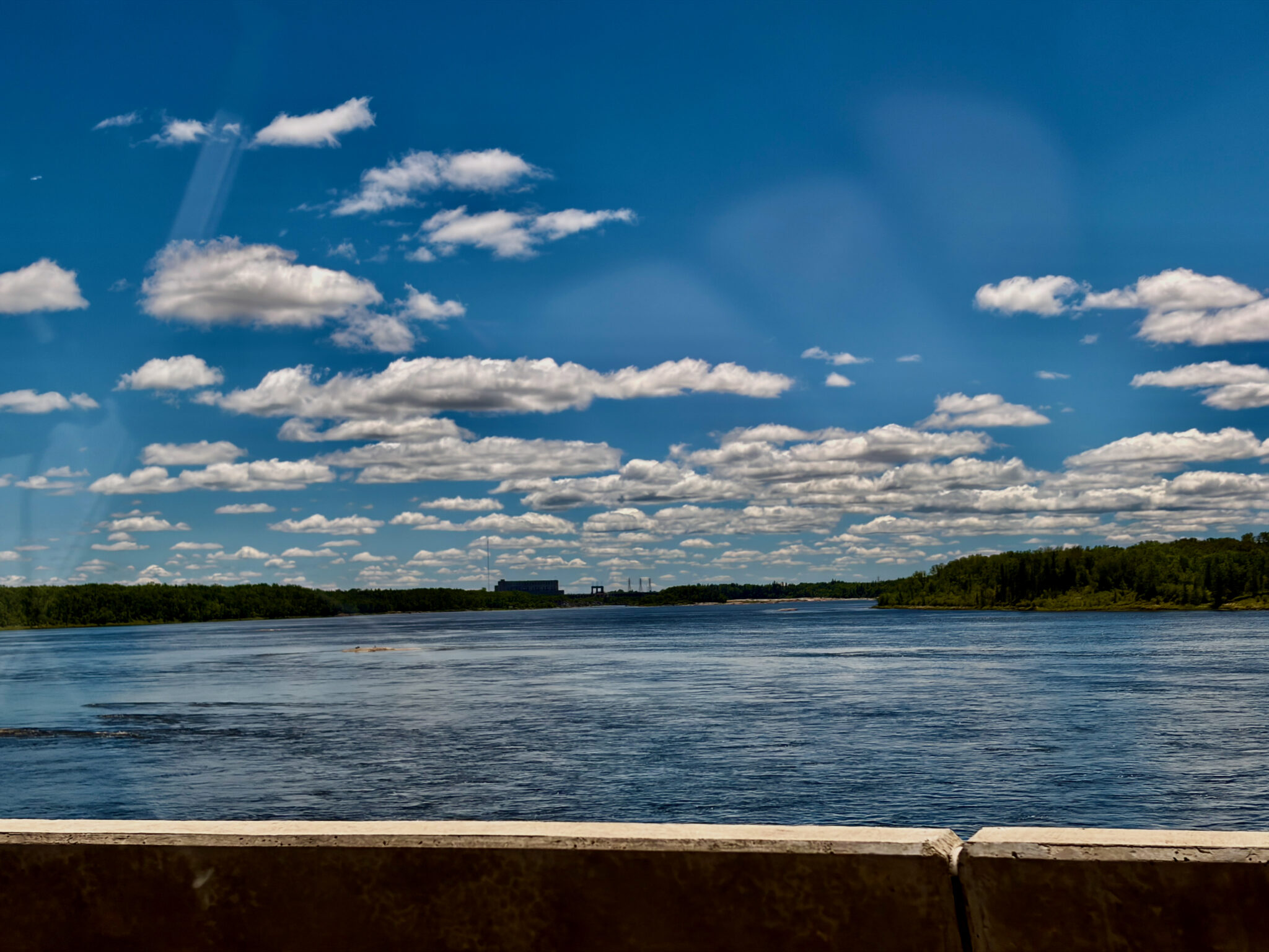 Wide river with forested shorelines under a bright blue sky dotted with white clouds, viewed from a concrete barrier in the foreground