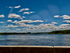 Wide river with forested shorelines under a bright blue sky dotted with white clouds, viewed from a concrete barrier in the foreground