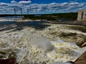 Turbulent river with high water levels and whitecaps flowing past three electrical transmission towers and a concrete structure, surrounded by forested shoreline under a partly cloudy sky