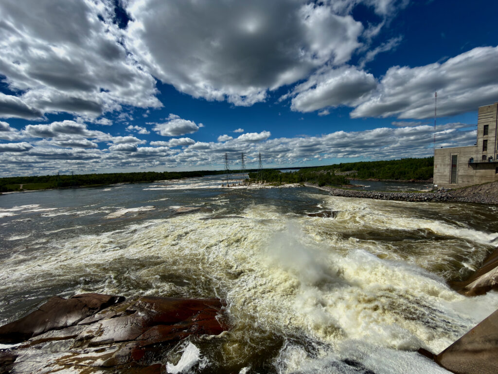 Turbulent river with white water rapids flowing past a concrete structure, with transmission towers and forested shoreline visible under a partly cloudy sky