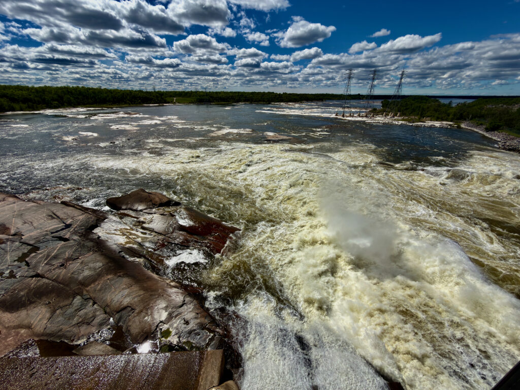 Turbulent river with rocky shoreline, forested banks, transmission towers in distance, and blue sky with white clouds