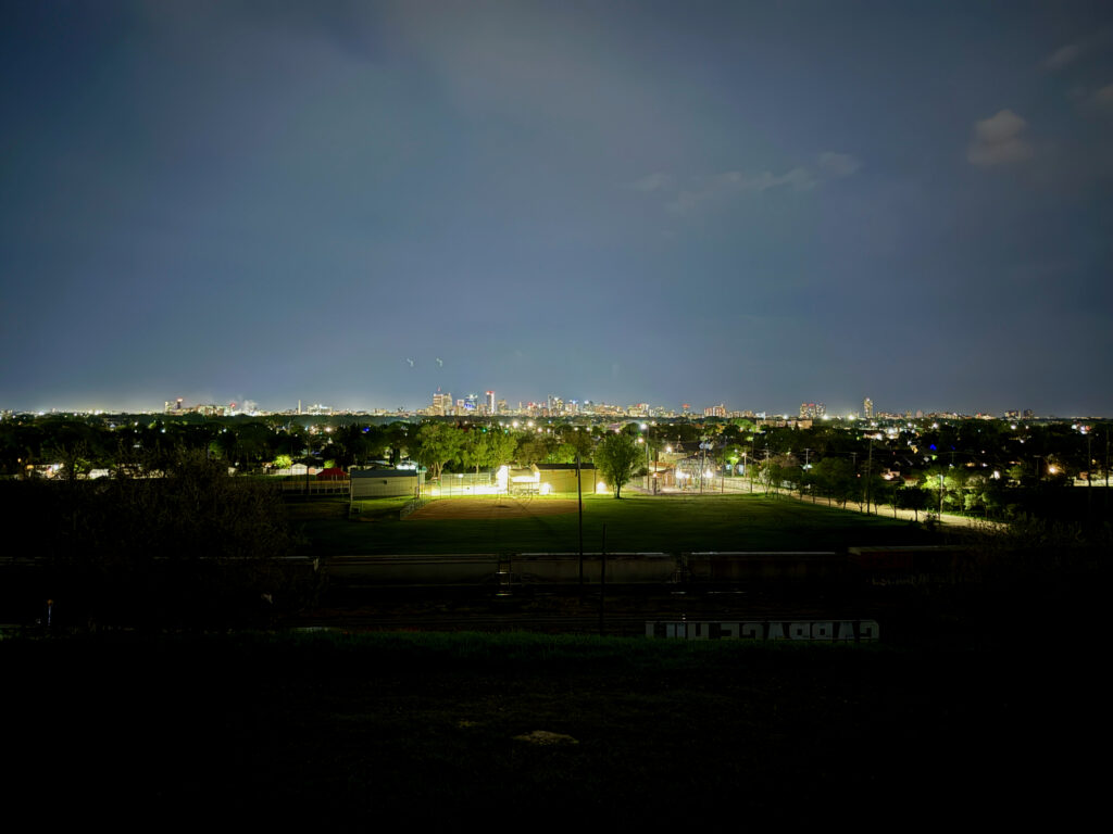 Westview Off-Leash Dog Park: Winnipeg's "Garbage Hill" and the First Park We Ever Walked in Canada 9 Distant city skyline at night with lights illuminating buildings and a foreground of trees and a sports field