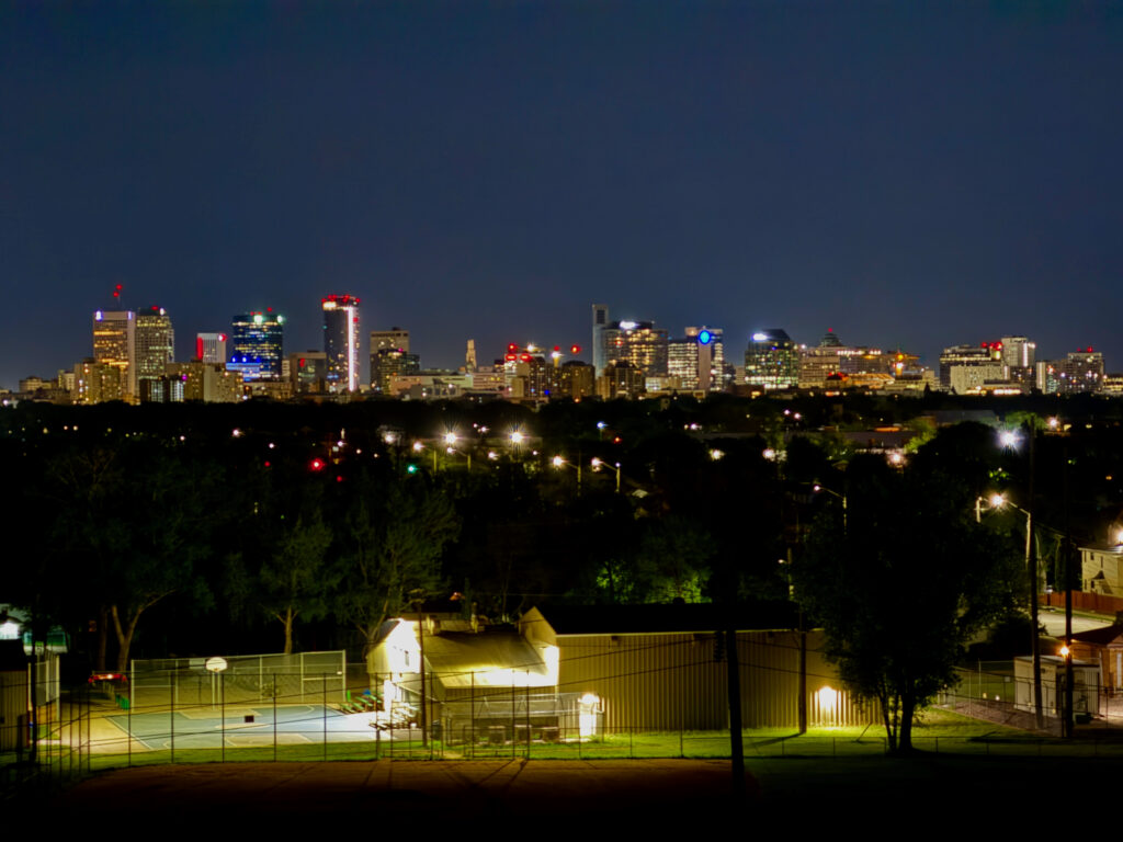 Westview Off-Leash Dog Park: Winnipeg's "Garbage Hill" and the First Park We Ever Walked in Canada 8 Nighttime view of a city skyline with illuminated buildings in the distance, overlooking a sports court and a brightly lit building in the foreground