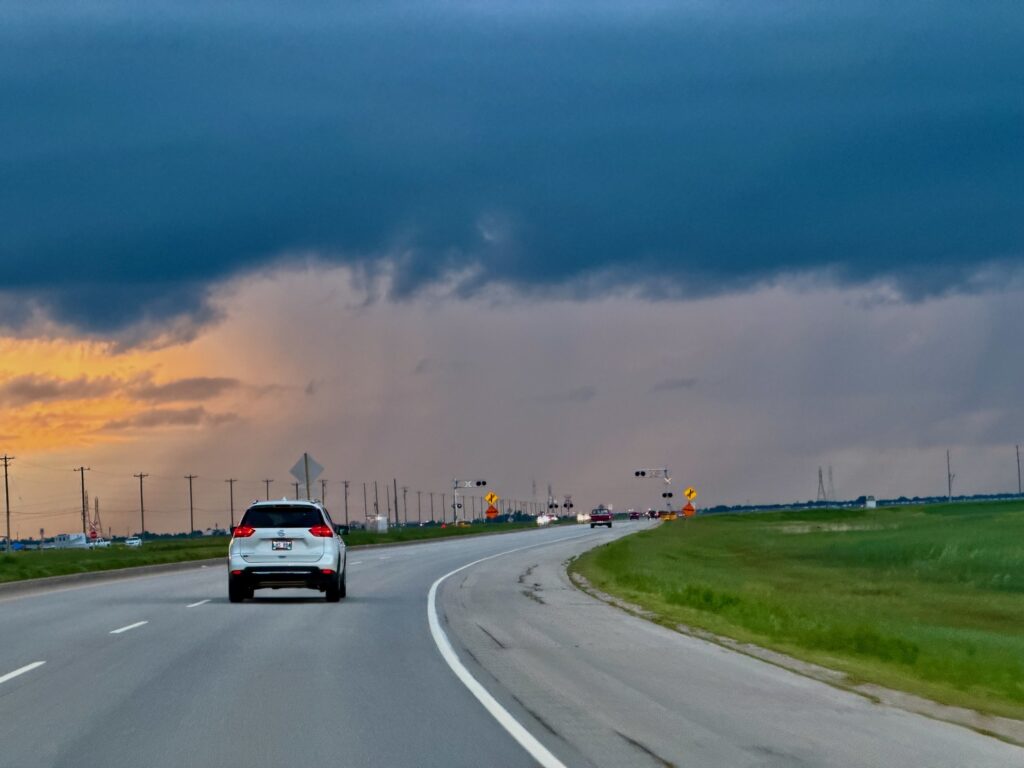 White car driving on a divided highway through flat farmland under a dramatic sky with dark clouds and golden sunset light