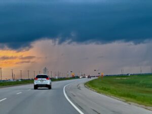 White car driving on a divided highway through flat farmland under a dramatic sky with dark clouds and golden sunset light