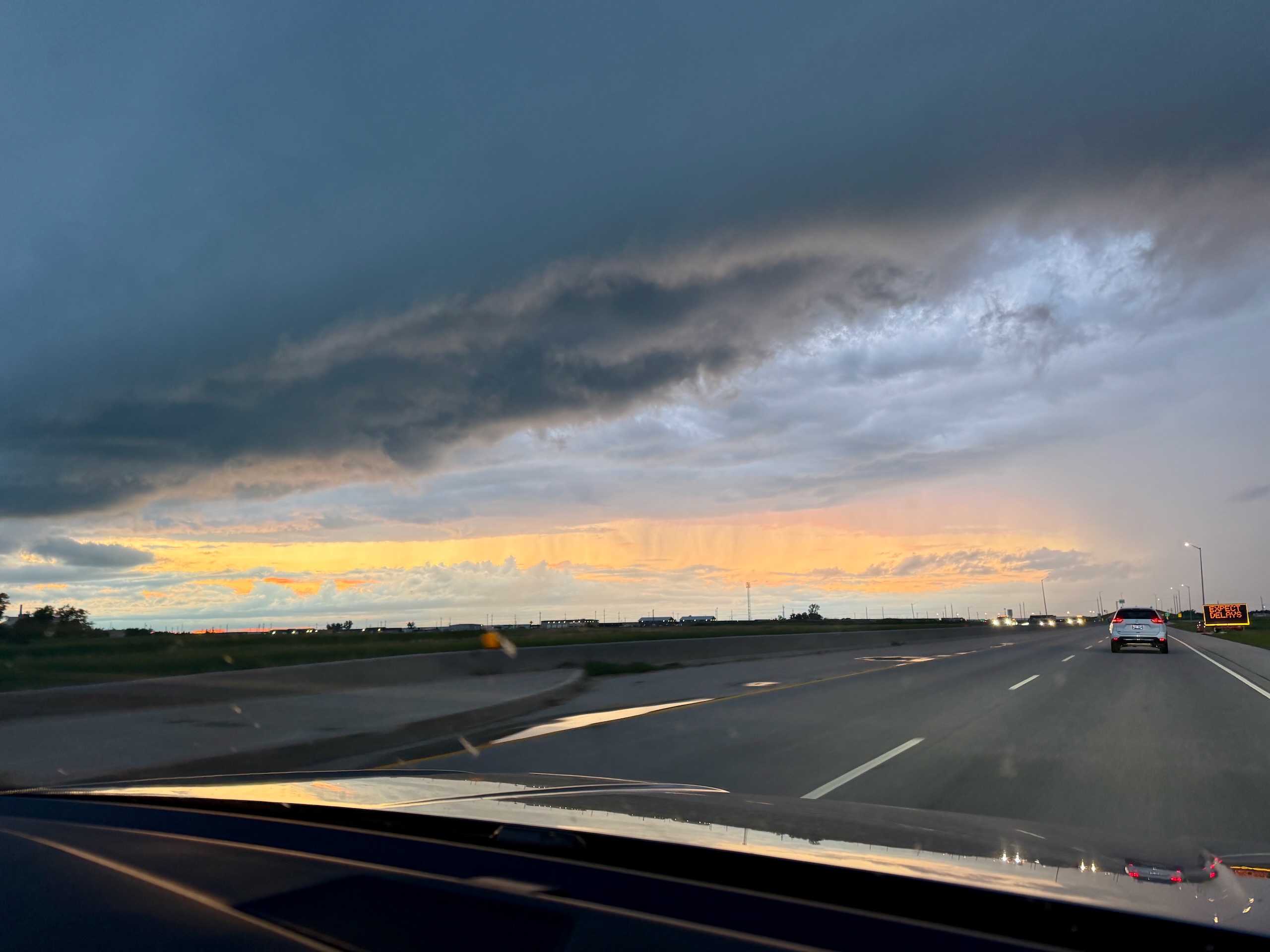 View from a vehicle on a highway during sunset, with dramatic clouds illuminated in yellow and orange against a blue sky, other vehicles ahead, and flat landscape with distant lights along the horizon