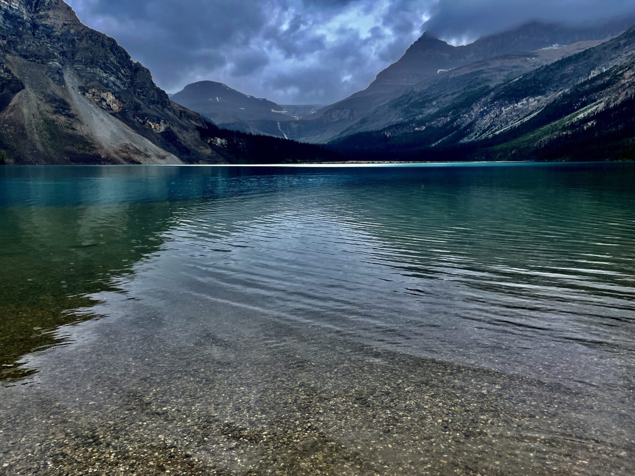 Turquoise alpine lake with clear shallow water and rocky shoreline, surrounded by steep forested mountains under a dramatic cloudy sky