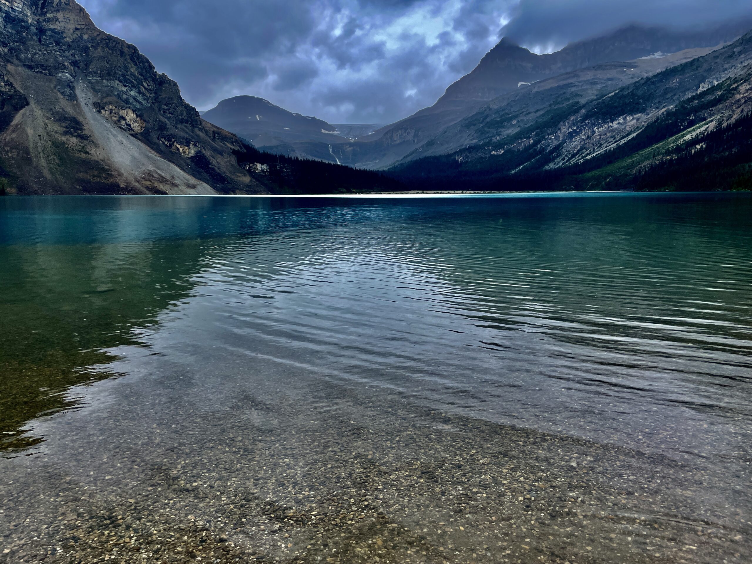 Turquoise alpine lake with clear shallow water and rocky shoreline, surrounded by steep forested mountains under a dramatic cloudy sky