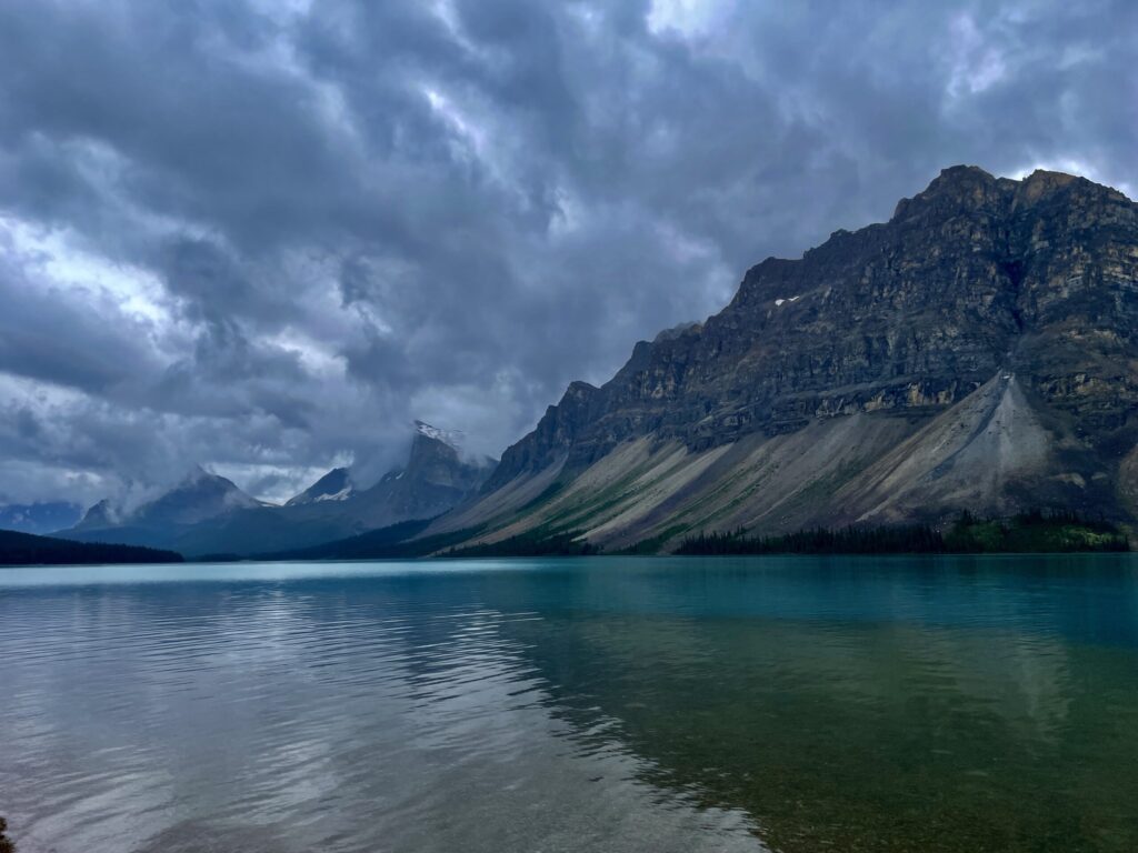 Turquoise alpine lake with dramatic rocky mountains and storm clouds overhead, coniferous forest along the shoreline