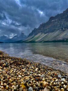 Rocky shoreline of a mountain lake with steep, striated peaks rising across the water under a dramatic cloudy sky