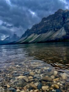 Clear shallow water with pebbles in foreground, mountain lake with forested slopes and dramatic cloudy sky, Canadian Rocky Mountains landscape