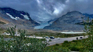Mountain valley with turquoise glacial lake, snow-capped peaks, and a highway with parked vehicles in the foreground under dramatic cloudy skies