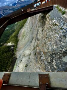 View from a mountain observation platform showing a steep rocky cliff face, forested valley below, and snow-capped peaks in the distance