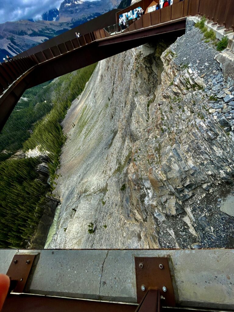 View from a mountain observation platform showing a steep rocky cliff face, forested valley below, and snow-capped peaks in the distance