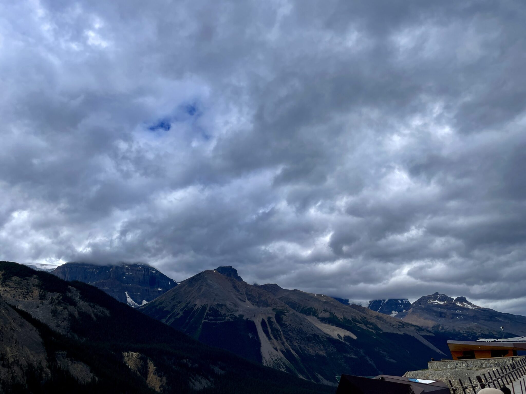 Dramatic cloudy sky over a mountain range with snow-capped peaks and forested slopes under overcast conditions