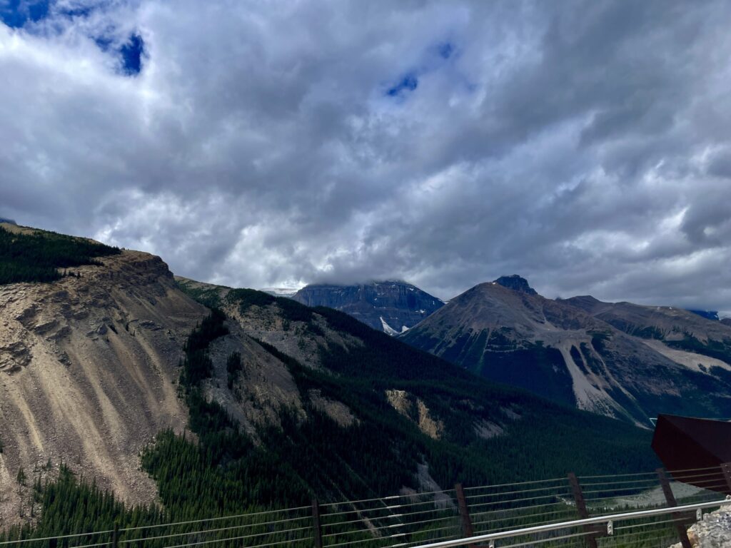 Mountain valley with forested slopes, rocky peaks, and dramatic cloudy sky viewed from a railed overlook