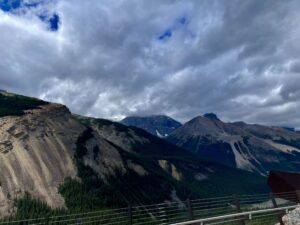 Mountain valley with forested slopes, rocky peaks, and dramatic cloudy sky viewed from a railed overlook