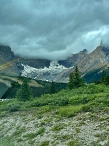 Alpine valley with glacier nestled between rocky peaks under stormy skies, coniferous forest in foreground, and patches of snow visible on mountainsides