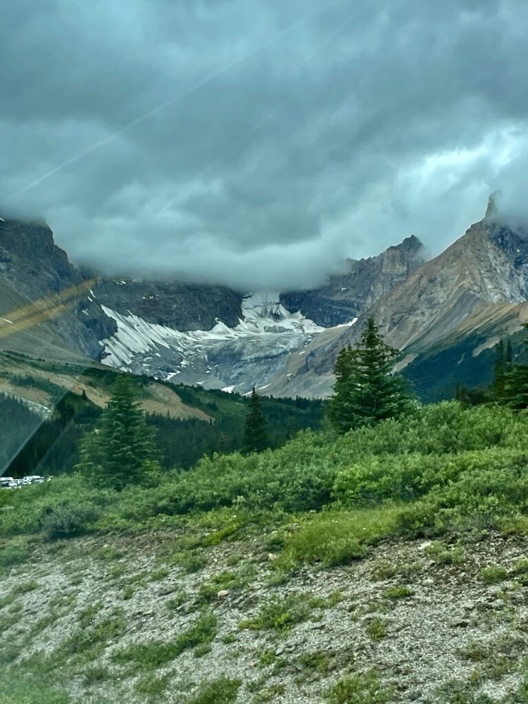 Alpine valley with glacier nestled between rocky peaks under stormy skies, coniferous forest in foreground, and patches of snow visible on mountainsides