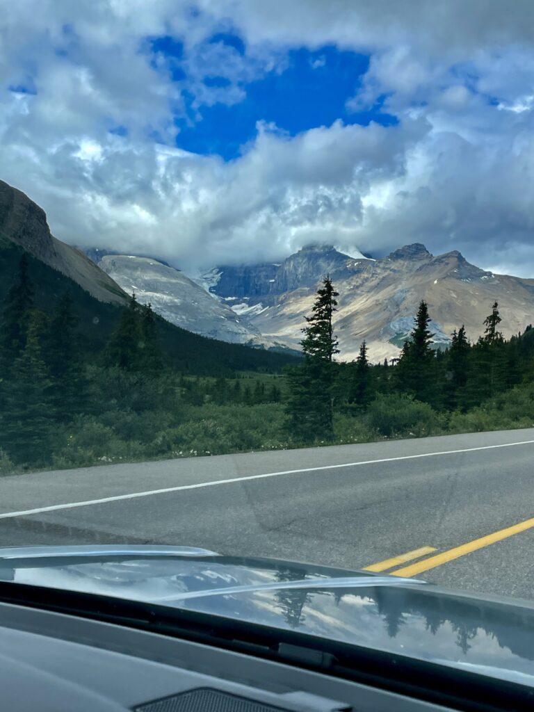 Mountain valley view from a vehicle windscreen showing snow-capped peaks, forested slopes, and a highway stretching through alpine terrain under partly cloudy skies