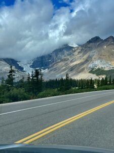 Mountain road view with coniferous forest, glacier-fed valley, and snow-capped peaks under partly cloudy sky
