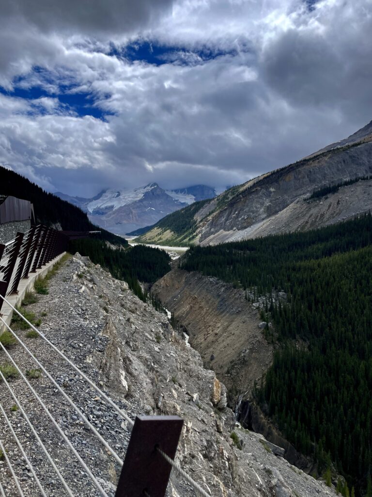 Mountain valley with snow-capped peaks, coniferous forest, and a glacial stream, viewed from a high rocky outcrop with metal railings under a partly cloudy sky